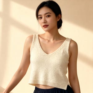 Könnte beinhalten: Cremefarbenes, ärmelloses Strickoberteil mit tiefem V-Ausschnitt. Das Oberteil hat einen gerippten Saum und eine leicht verkürzte Länge. Der Stoff wirkt weich und strukturiert. Das Model trägt das Oberteil mit dunkelblauen Hosen.