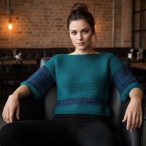 May include: Teal and navy blue crochet sweater with a boat neck and three-quarter sleeves. The sweater has a horizontal navy blue stripe across the midsection. The model is wearing black pants and is seated in a black chair.