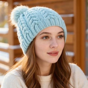 Può includere: Un berretto lavorato a trecce blu chiaro con un soffice pompon. Il cappello è realizzato in un materiale morbido e strutturato. Il berretto è un accessorio invernale, perfetto per il freddo. Il cappello è indossato da una persona.