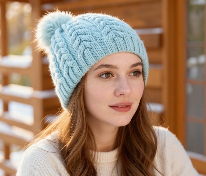 May include: A light blue cable-knit beanie with a fluffy pom-pom. The hat is made of a soft, textured material. The beanie is a winter accessory, perfect for cold weather. The hat is worn by a person.