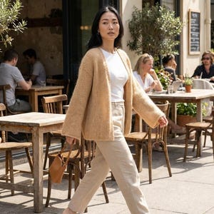 May include: A woman wearing a tan cardigan, white top, and beige pants walks on a sidewalk. She carries a small brown purse. The background shows a cafe with outdoor seating and other people.