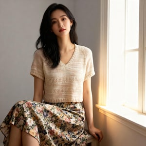 以下が含まれることがあります： ベージュのVネックニットトップス、半袖。胸元に水平模様のデザイン。モデルは花柄のスカートを着用しています。カジュアルウェアに適した服装です。