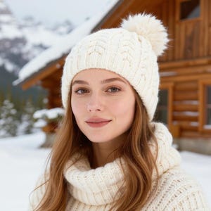 May include: Cream-colored cable knit hat with a fluffy pom-pom. The hat is worn with a matching cream-colored sweater. The background shows a snow-covered landscape and a wooden cabin.