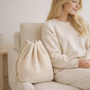 Può includere: Una borsa a coulisse all'uncinetto color crema. La borsa è realizzata in filato spesso, con un motivo a punto strutturato. La borsa ha una forma arrotondata e una chiusura con coulisse. La borsa è su un divano color crema.