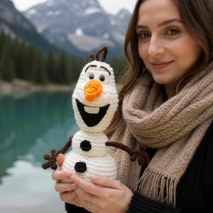 May include: A crocheted Olaf snowman doll with a carrot nose, black coal buttons, and twig arms. The snowman is white with a black smile and is held in front of a scenic lake and mountain backdrop.