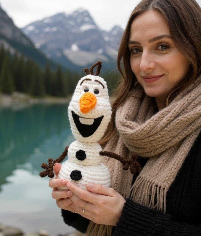 May include: A crocheted Olaf snowman doll with a carrot nose, black coal buttons, and twig arms. The snowman is white with a black smile and is held in front of a scenic lake and mountain backdrop.