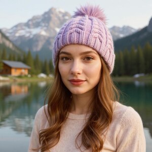 May include: A light purple cable-knit beanie with a matching pom-pom. The hat is worn by a person with long brown hair, a light pink sweater, and a scenic mountain and lake backdrop.
