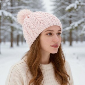 May include: A light pink cable-knit beanie with a matching pom-pom. The hat is worn by a person in a cream-colored sweater, set against a snowy forest backdrop. The beanie's texture and color create a cozy winter aesthetic.
