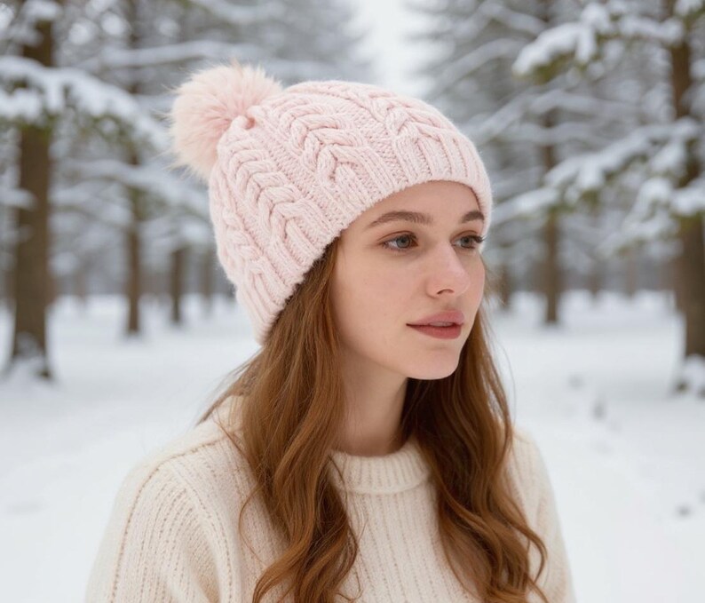 May include: A light pink cable-knit beanie with a matching pom-pom. The hat is worn by a person in a cream-colored sweater, set against a snowy forest backdrop. The beanie's texture and color create a cozy winter aesthetic.