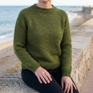 May include: Olive green knit sweater with a crew neck and long sleeves. The sweater has ribbed cuffs and hem. The person is wearing black pants. The background shows a beach and ocean.