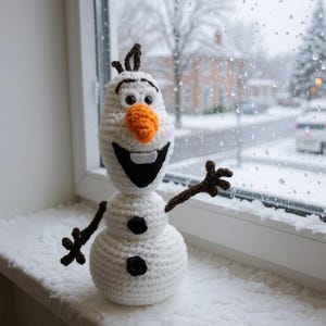 May include: A crocheted snowman with a carrot nose, black coal buttons, and twig arms. The snowman is white with a black smile and eyebrows. It is sitting on a windowsill with a snowy background.