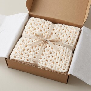 May include: Cream-colored, crocheted blanket tied with a beige ribbon, presented in a brown cardboard box. The blanket has a textured, open weave design. The box is lined with white tissue paper, creating a gift-like presentation.