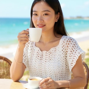 Op de afbeelding: Een witte gehaakte top met een ronde hals. Een witte koffiekop wordt dicht bij het gezicht gehouden. Een tweede witte kop koffie staat op een schotel op een houten tafel. De achtergrond toont een strand en de oceaan.