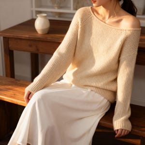 May include: A cream-colored sweater with an off-the-shoulder neckline. The long-sleeved knitwear is paired with a white skirt. A small, off-white vase sits on a wooden table in the background. The sweater has a ribbed texture on the cuffs and hem.