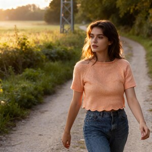 May include: A peach-colored, short-sleeved knit top with a scalloped hem. The top has a round neckline and a small cutout detail on the shoulder. The person is wearing blue jeans and walking on a dirt path.