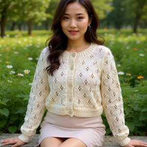 May include: A cream-coloured, open-knit cardigan with a round neckline and button closure. The long-sleeved sweater features a delicate diamond pattern and is paired with a light pink skirt. The image is set outdoors with a blurred background of greenery and flowers.