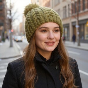 May include: A green cable-knit beanie with a faux fur pom-pom. The hat is worn by a person wearing a black coat. The background shows a city street with buildings and a crosswalk.