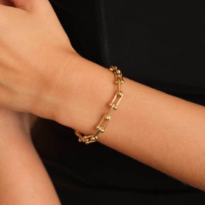 Puede incluir: Un brazalete dorado con un diseño de eslabones de cadena. El brazalete está hecho de pequeños eslabones redondeados y se lleva en la muñeca sobre un fondo negro. La joya es un accesorio de moda.