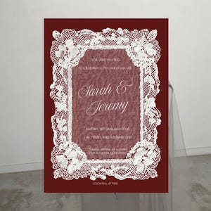 May include: A burgundy wedding invitation with white lace trim and text. The invitation reads "Sarah & Jeremy" and includes the date, time, and location of the ceremony. The invitation is for cocktail attire.