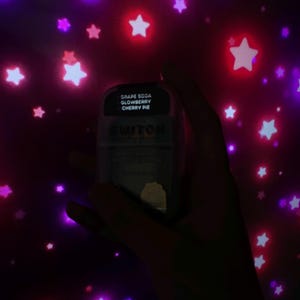May include: A clear, rectangular product with the text "GRAPE SODA GLOWBERRY CHERRY PIE" is held against a background of glowing purple and red stars. The product has the word "SWITCH" printed on it.