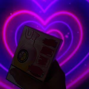 May include: A box with a pink and yellow design, held against a background of neon purple hearts. The box features the letters "UT" and a graphic of a bottle with a lemon slice. The background is a gradient of purple and pink.