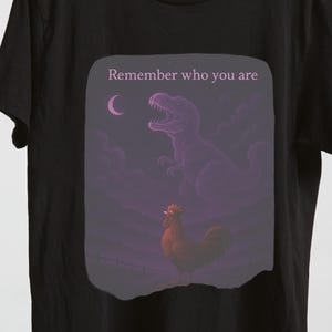Puede incluir: Camiseta negra con un diseño gráfico que presenta un dinosaurio y un gallo bajo una luna creciente. El texto "Remember who you are" está encima de la imagen. La camiseta está colgada en una percha de madera.