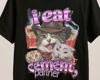 May include: Black t-shirt featuring a graphic of a winking cat wearing a cowboy hat, with the text "i eat cement partner" in pink. The design includes two other cat faces and lightning bolt accents.