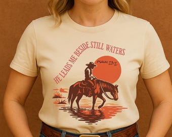 Könnte beinhalten: Beiges T-Shirt mit einem Cowboy auf einem Pferd in Wasser, mit roter Sonne im Hintergrund. Der Text "HE LEADS ME BESIDE STILL WATERS" und "PSALM 23:2" ist auf dem Shirt aufgedruckt.