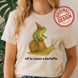 Camiseta Cottagecore con rana montando caracol: divertida camiseta gráfica retro con la frase "A causar un alboroto".