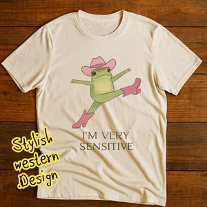 May include: A cream-coloured t-shirt with a cartoon frog wearing a pink cowboy hat and boots, with arms and legs outstretched. The text "I'M VERY SENSITIVE" is below the frog. The words "Stylish western Design" are in a yellow font on the left side of the shirt.