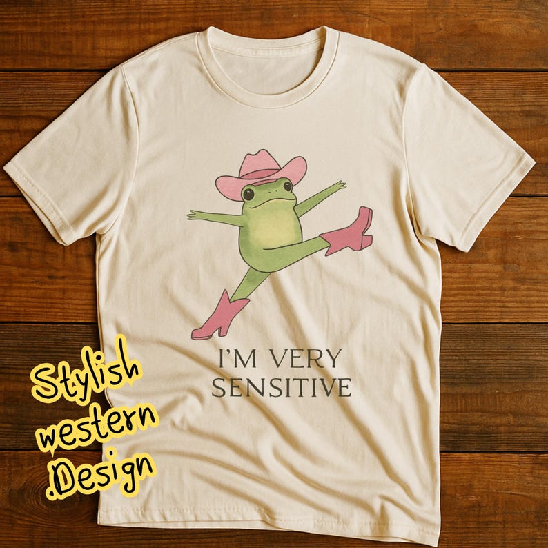 May include: A cream-coloured t-shirt with a cartoon frog wearing a pink cowboy hat and boots, with arms and legs outstretched. The text "I'M VERY SENSITIVE" is below the frog. The words "Stylish western Design" are in a yellow font on the left side of the shirt.