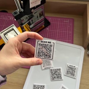 Peut inclure: Un aimant carré blanc avec un code QR noir et le texte "SCAN ME" est tenu par une main. Plusieurs aimants similaires sont sur une surface blanche. Une machine à fabriquer des boutons noire et argentée est en arrière-plan.