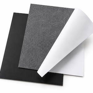 May include: A stack of three rectangular sheets of material. The bottom sheet is solid black, the middle sheet is a textured dark grey felt, and the top sheet is white with a corner folded over to reveal a smooth, bright white surface texture.