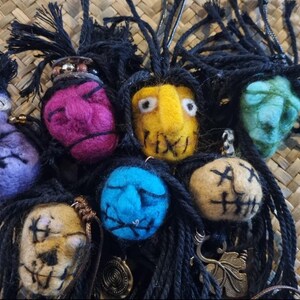 Puede incluir: Una colección de colgantes de fieltro hechos a mano con caras estilizadas en varios colores, incluyendo morado, amarillo, azul y verde. Cada colgante tiene pelo de hilo negro y una expresión facial cosida. Algunos colgantes tienen abalorios metálicos.