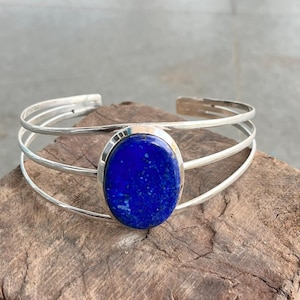 May include: A silver cuff bracelet featuring a large, oval, deep blue stone. The bracelet has three parallel bands. The stone is set in a silver bezel. The bracelet is displayed on a wooden surface.