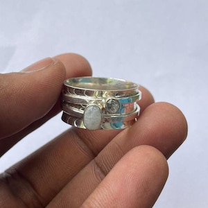 Peut inclure: Une bague spinner argentée avec trois bandes et deux pierres précieuses. Une pierre précieuse est de forme ovale et l'autre est ronde. La bague est tenue dans la main d'une personne.