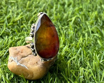 Op de afbeelding: Een zilveren ring met een grote, druppelvormige edelsteen. De edelsteen heeft een mix van kleuren, waaronder bruin, oranje en rood. De ring is geplaatst op een kleine, bruin gekleurde steen, met een achtergrond van groen gras.