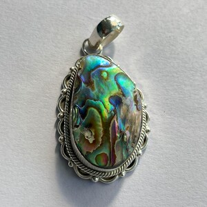 May include: Abalone shell pendant with a silver bail. The iridescent shell displays a mix of green, blue, and purple hues. The pendant is set in a silver frame with a decorative rope-like border.