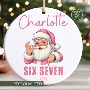 May include: A white, round Christmas ornament featuring a cartoon Santa Claus in a pink suit and hat. The ornament is personalized with the name "Charlotte" and the text "SIX SEVEN 2025". A gold string is attached for hanging.