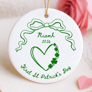 May include: White ceramic ornament with a green bow, heart, and shamrock design. The ornament has the name "Niamh" and the year "2026" printed on it, along with the text "First St Patrick's Day". A gold string is attached for hanging.
