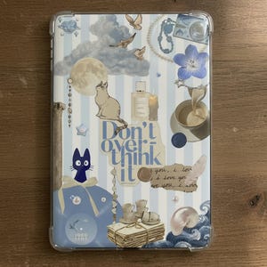 May include: Clear Kindle case with a collage design featuring a cat, moon, clouds, and the text "Don't overthink it." The case has a blue and white colour scheme with decorative elements like flowers and birds.