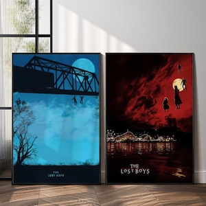 May include: Two framed art prints. The left print features a bridge silhouette against a blue sky with figures falling. The right print shows figures against a red sky with a full moon and a waterfront scene. Both prints have "THE LOST BOYS" text.