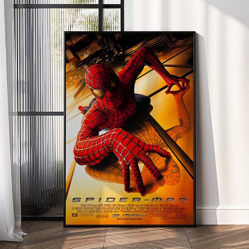 May include: Framed movie poster featuring Spider-Man in a red and blue suit, with a cityscape background. The poster has the title "Spider-Man" in bold white letters. The poster is displayed indoors, leaning against a wall.
