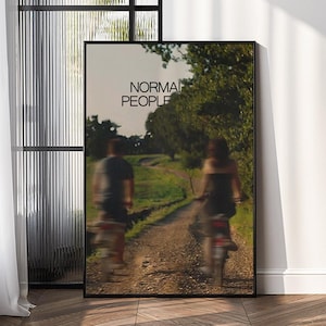 May include: A framed art print featuring the text "NORMAL PEOPLE" at the top. The image shows two blurred figures riding scooters on a dirt path, with trees lining the sides. The artwork is displayed against a white wall.