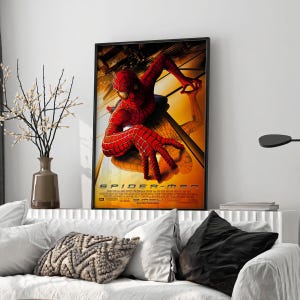 May include: Framed movie poster for Spider-Man, featuring the superhero in a red and blue suit, clinging to a building. The poster has a yellow and orange background with the movie title and credits at the bottom. The poster is displayed above a white couch.