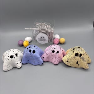 Puede incluir: Cuatro figuras de cerámica con forma de fantasma moteado en blanco, azul, rosa y amarillo. Una pequeña bolsa con relleno blanco y unos cuantos huevos moteados están detrás de las figuras. Las figuras tienen tres agujeros negros para ojos y boca.