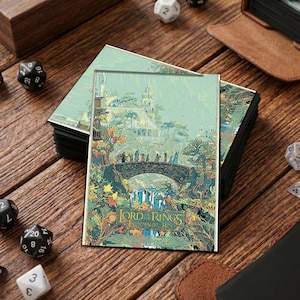 May include: A stack of fantasy art cards featuring a bridge scene with a waterfall and a castle in the background. The cards have the text "LORD OF THE RINGS FELLOWSHIP OF THE RING" at the bottom. Dice and a leather-bound book are also visible.