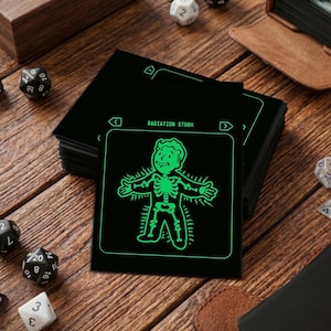 Op de afbeelding: Zwarte kaart met een groene illustratie van een skelet en de tekst "RADIATION STORM". De kaart maakt deel uit van een stapel op een houten oppervlak, met dobbelstenen en een lederen item op de achtergrond.