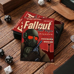 May include: A stack of Fallout: New Vegas game cards featuring a soldier in a gas mask holding a gun. The cards have a red background with the game title and text. Dice and a leather-bound book are in the background.