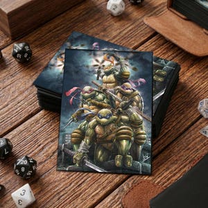 Op de afbeelding: Een stapel kaarthoezen met de Teenage Mutant Ninja Turtles. De illustratie toont de vier schildpadden in actie, met getrokken wapens, tegen een donkere, bewolkte achtergrond. Dobbelstenen en andere spelaccessoires liggen verspreid.